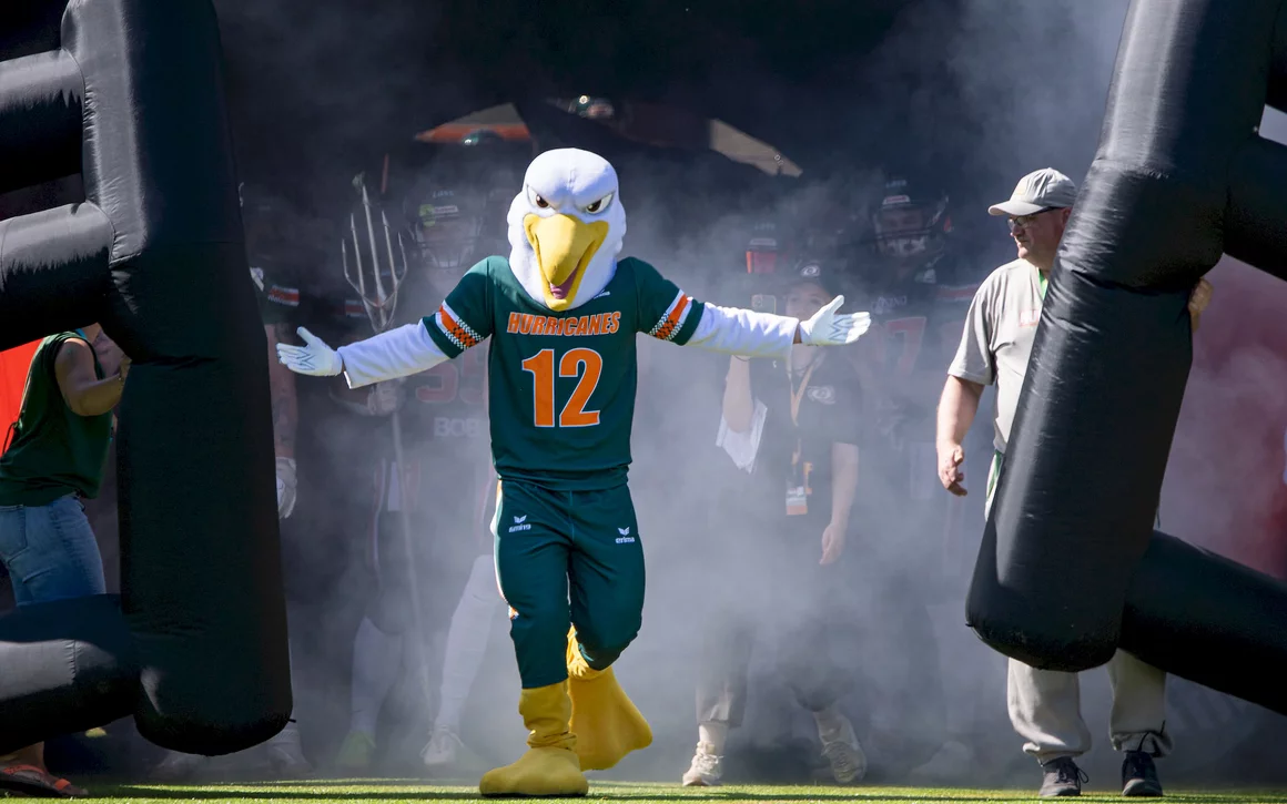Das Maskottchen der Baltic Hurricanes, ein Vogel in grün-orangefarbener Football-Ausrüstung mit der Nummer 12, läuft durch einen aufblasbaren Tunnel auf das Spielfeld. Rauch- oder Nebeleffekte sind hinter ihm zu sehen