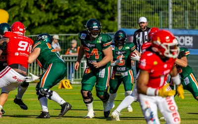 „Ein American-Football-Spiel auf einem grünen Spielfeld. Mehrere Spieler von zwei Teams, in roten und grünen Tricots laufen mit dem Ball, die Spieler verfolgen sich und versuchen zu blockieren. Im Hintergrund stehen Zuschauer.