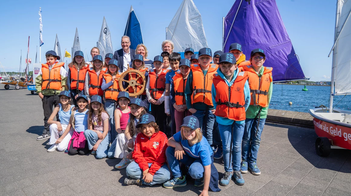 Zur Eröffnung des Segelcamps überreichen Bettina Aust, Dr. Jörg Teupen und Uwe Wanger das Steuerrad an die Segelkinder der Friedrich-Junge-Grundschule. (Foto: Kiel-Marketing e.V.)