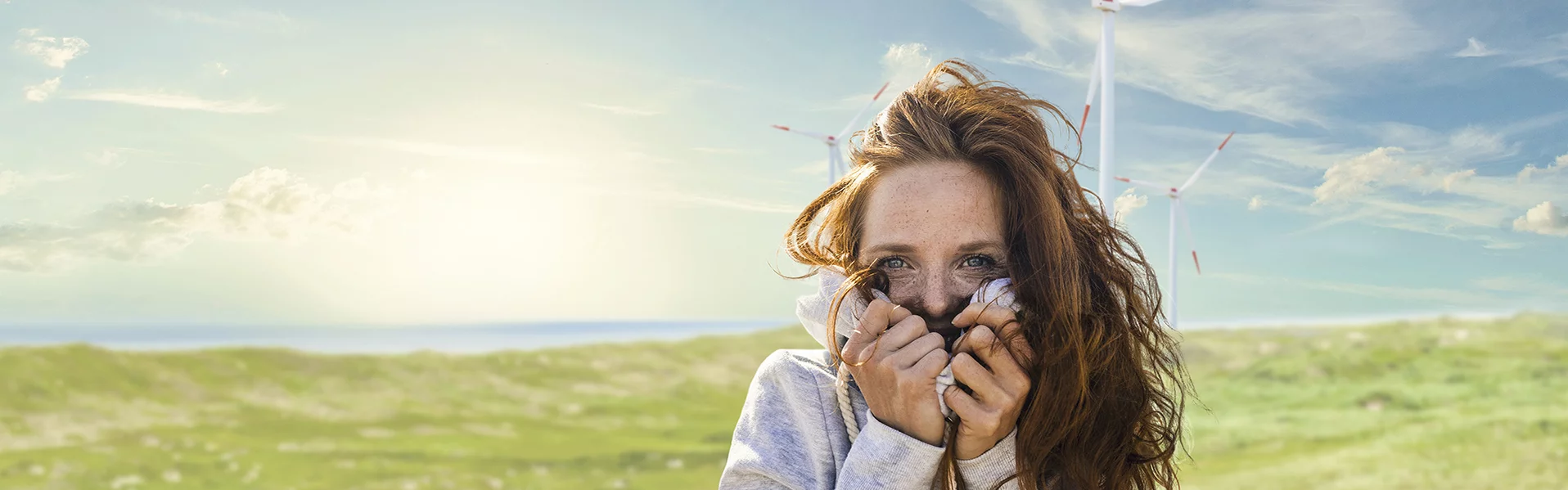 Sich in ihren Pullover kuschelnde, lächelnde Frau auf einer Wiese vor einem Windrad