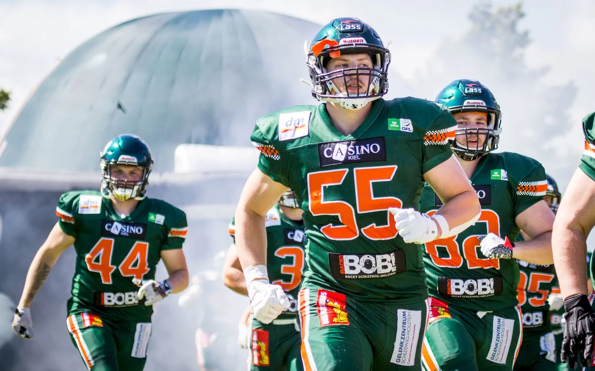 Eine Gruppe von American-Football-Spielern in grünen Trikots mit orange-weißen Akzenten läuft aus dem Tunnel auf das Spielfeld. Im Vordergrund ist ein Spieler mit der Nummer 55 gut sichtbar. Im Hintergrund sind eine große Kuppelstruktur und Nebel oder Rauch zu sehen.