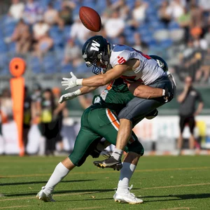 Zwei American-Football-Spieler sind mitten im Spielgeschehen zu sehen. Ein Spieler im grünen Trikot scheint einen Spieler im blauen Trikot zu tackeln oder zu blocken. Im Hintergrund sind Tribünen und einige Zuschauer zu erkennen